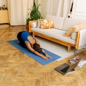 Woman doing yoga indoors on a mat with a laptop nearby, promoting wellness and fitness.