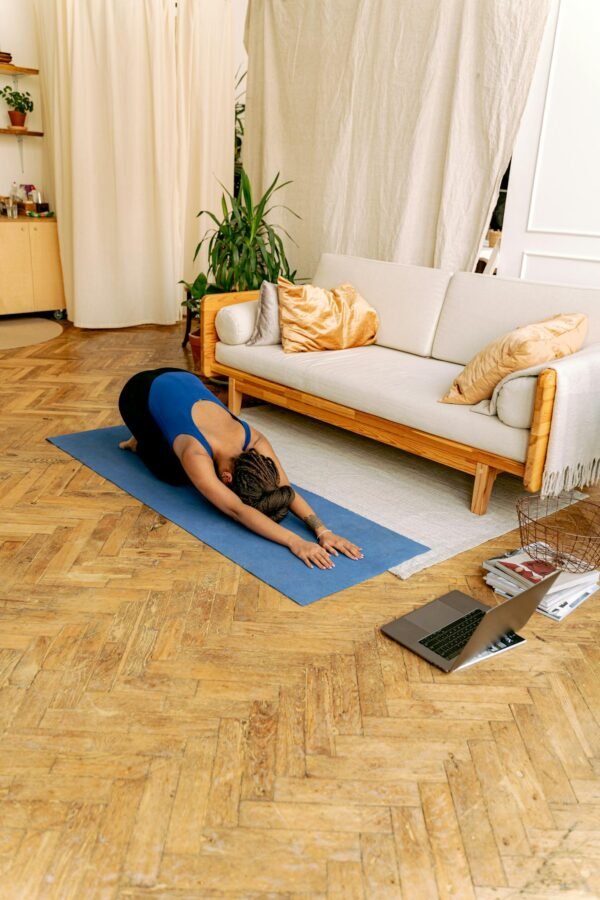 Woman doing yoga indoors on a mat with a laptop nearby, promoting wellness and fitness.
